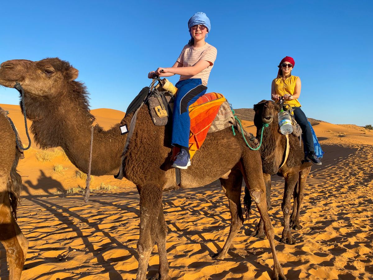 Son and mom atop dromedaries in the Saharan Desert in Morocco